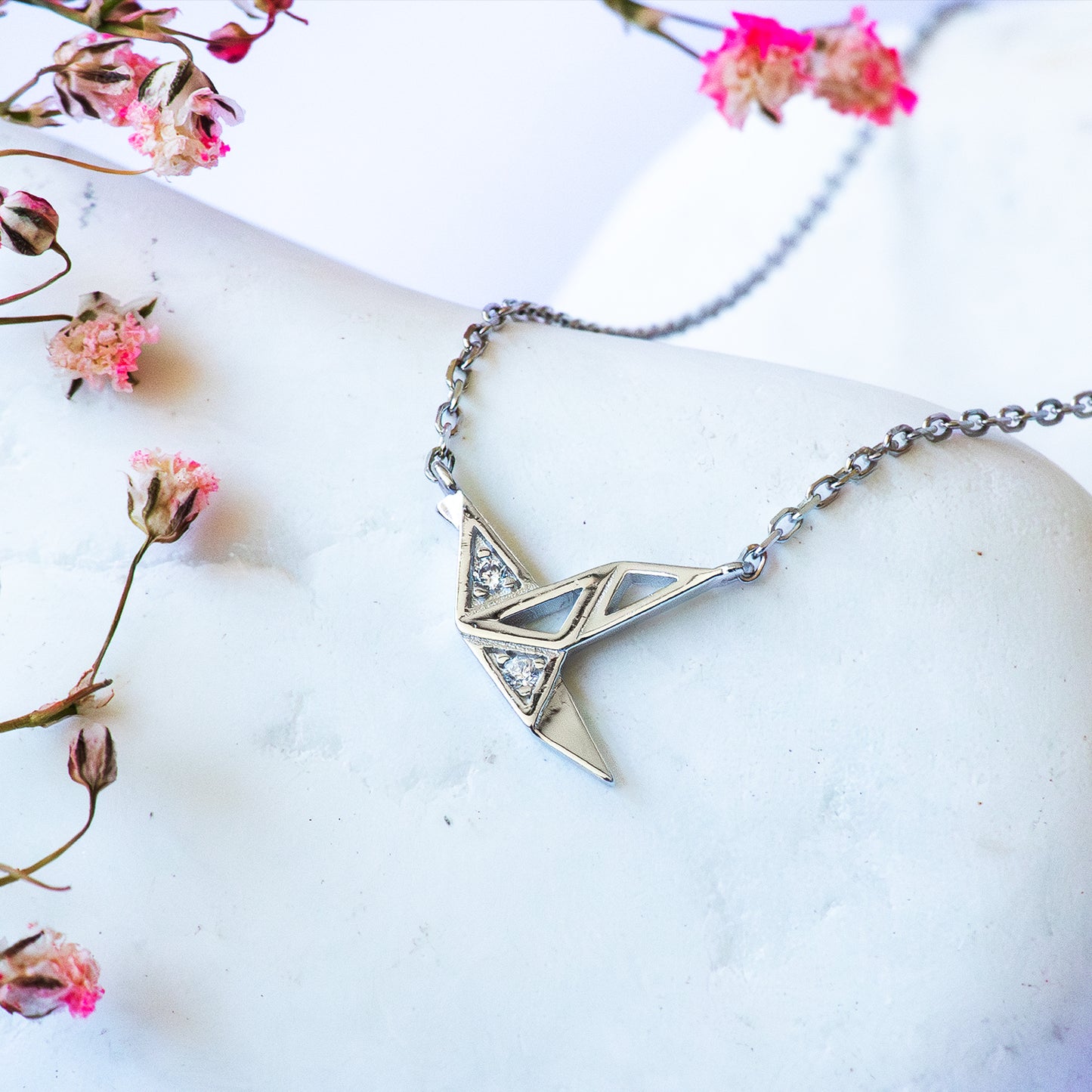 Colgante y cadena con grulla tipo origami adornada con circones sobre piedra blanca con flores