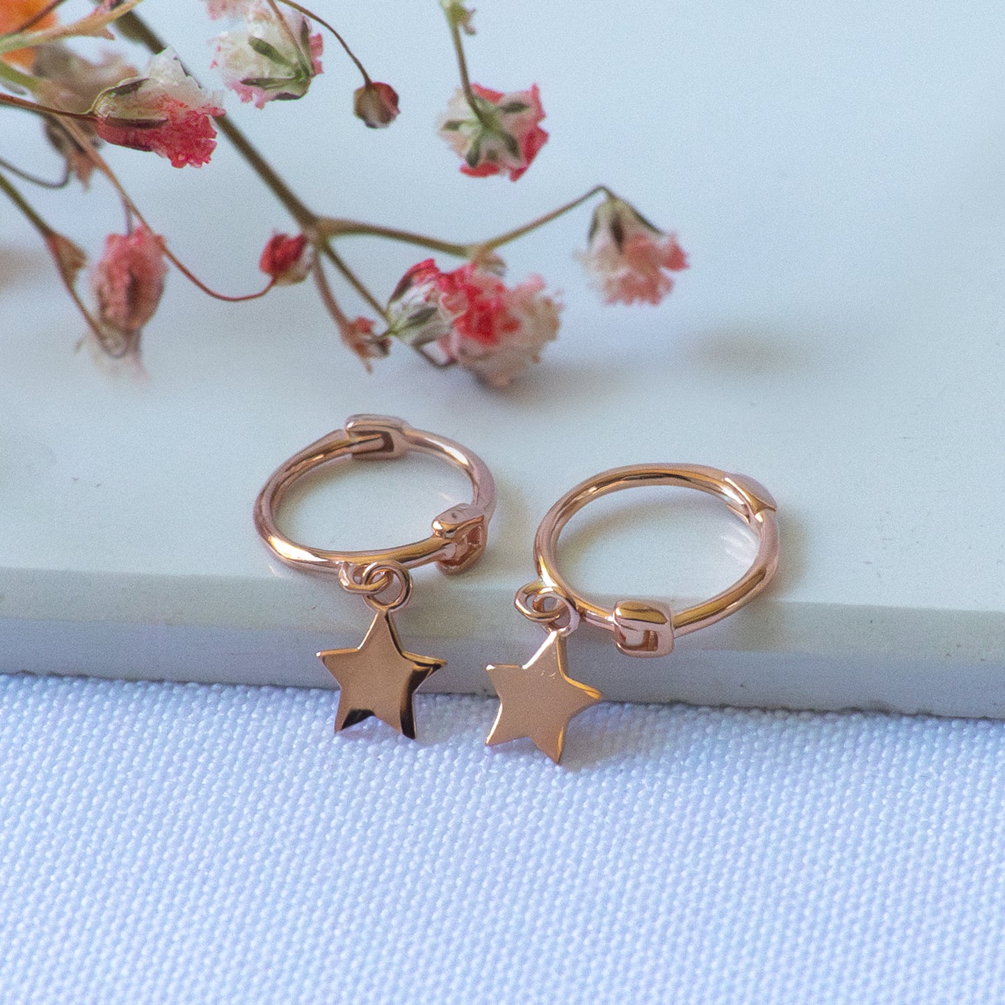 Par de aros de oro rosado con colgante de estrella plana sobre piedra con flores.