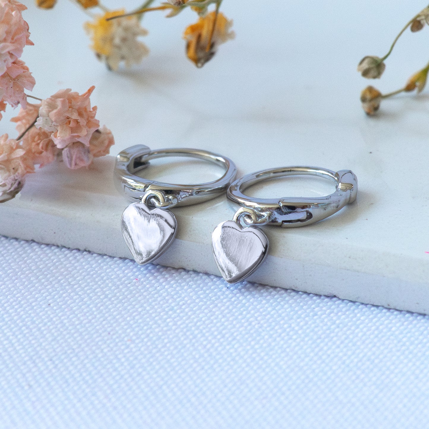 Par de aros plateados con colgante de corazón sobre superficie blanca.