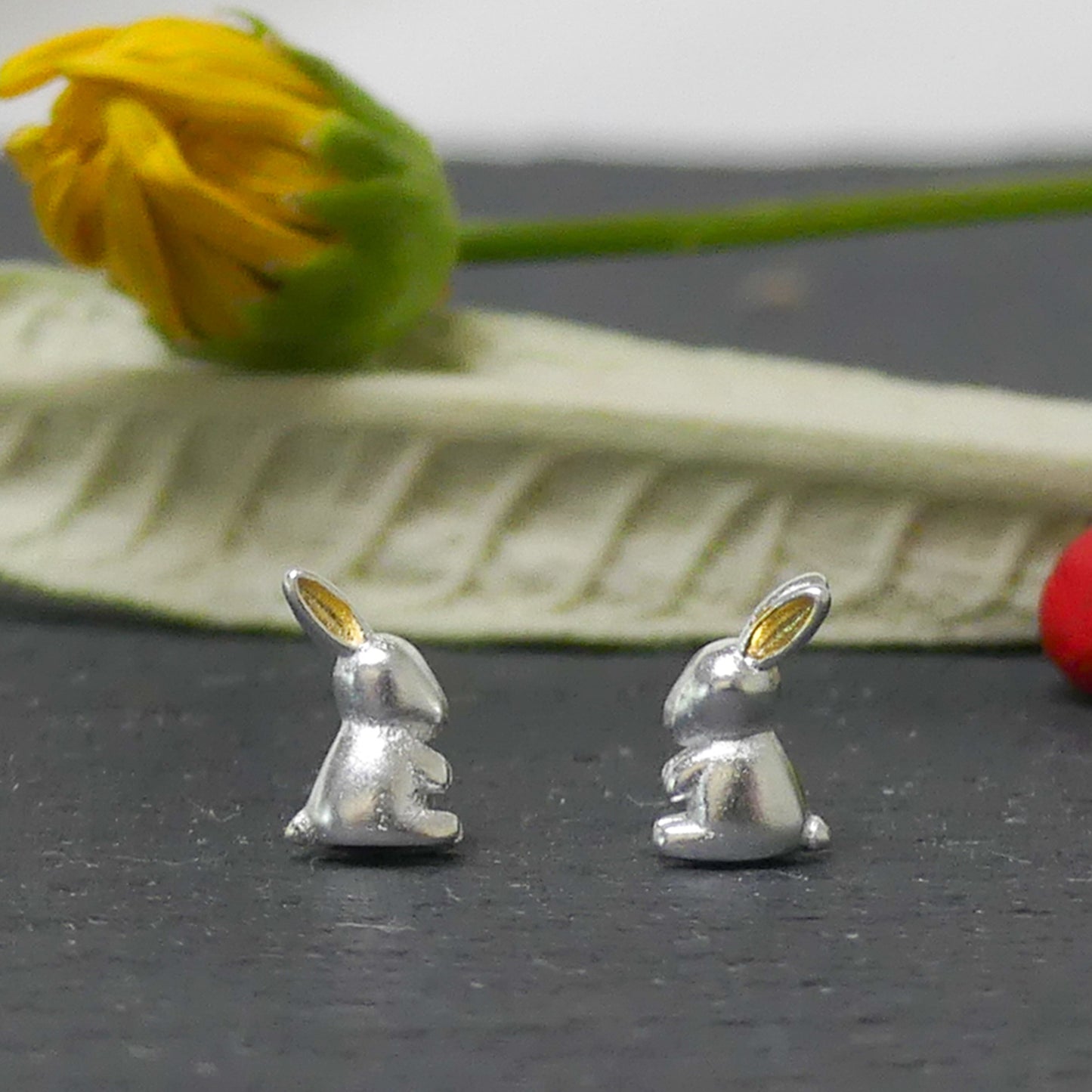 Aritos de plata y oro, en en forma de conejito de perfil plateado con orejas doradas sobre piedra gris y flor amarilla