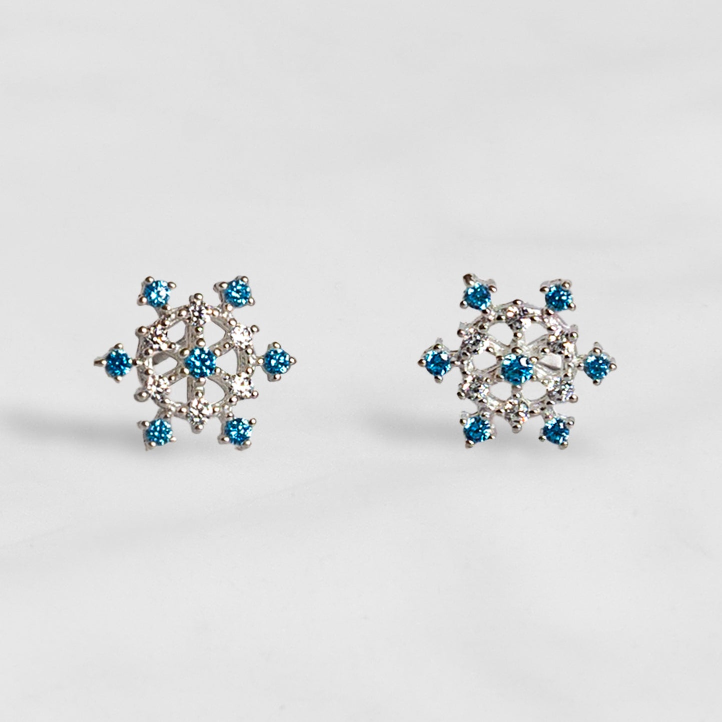Par de aros de plata en forma de copo de nieve con circones blancos y azules sobre fondo gris