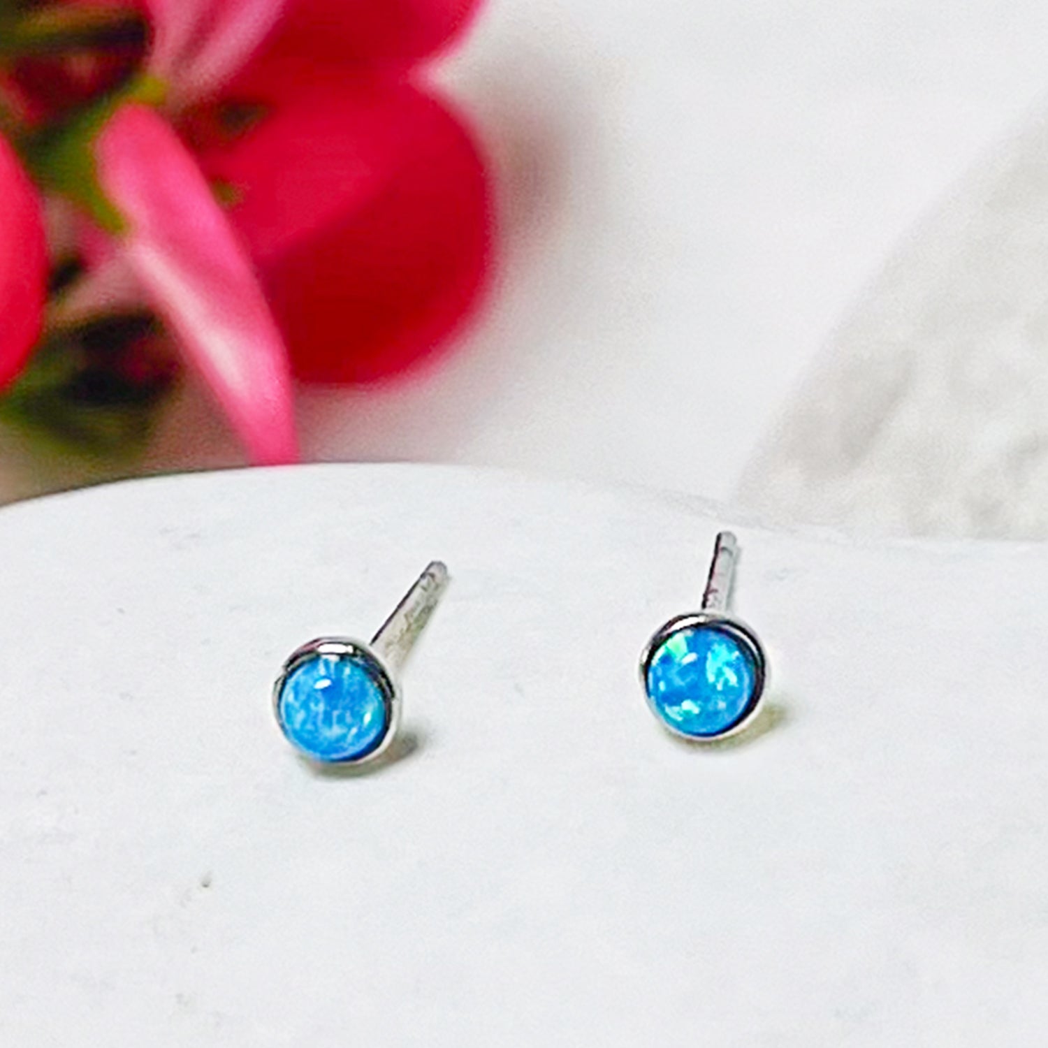 Par de aritos con ópalo azul sobre superficie blanca con flores rosadas difuminadas