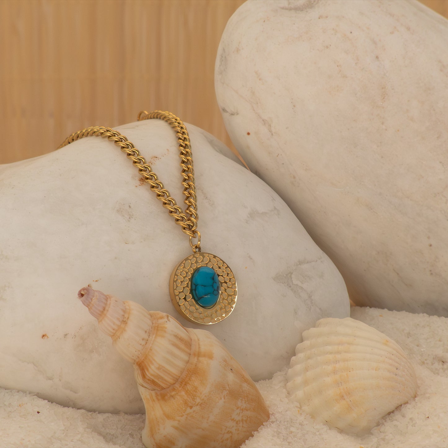 Collar boho con disco dorado y piedra turquesa sobre arena con conchas