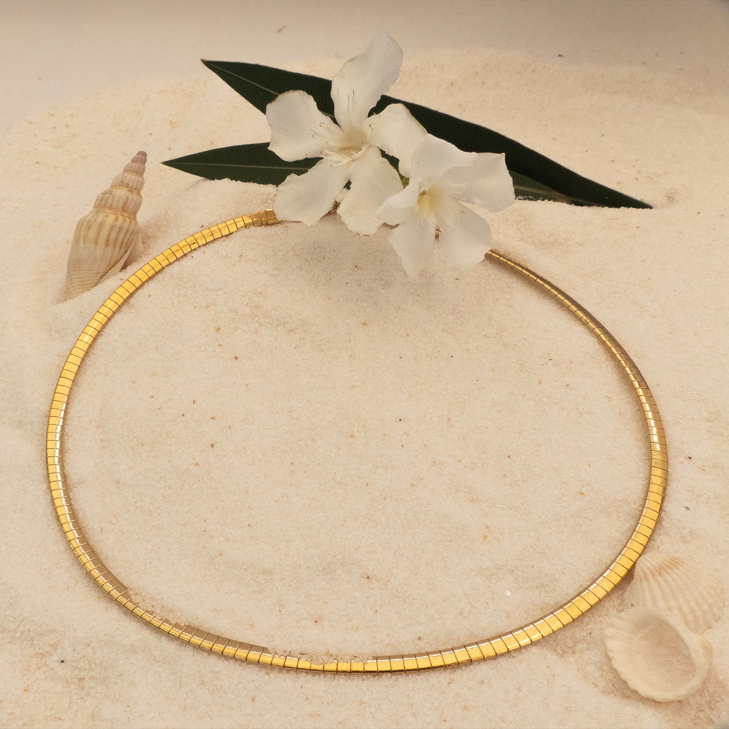 Collar dorado tipo choker rígido sobre arena con conchas y flor blanca